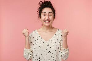 Glad young curly brunette girl with brown eyes in white blouse with polka dots raised hands and clenched fists in excitement when got the victory and became a winner in competition, over pink wall photo