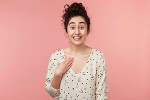 Young cheerful attractive curly brunette girl with brown eyes in white blouse with polka dots pointing with hand at herself in excitement being chosen for the main role in university play photo