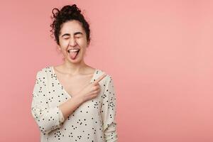 Curly young brunette girl in white blouse with polka dots has fun, closed eyes and shows tongue while pointing to the upper right corner with index finger on empty copy space, over pink background photo