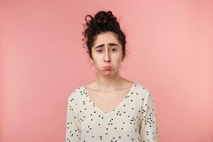 Brunette girl offended sad upset, pouted her cheeks is overwhelmed with negative emotions, in bad mood dressed in blouse with polka dots, going to cry, isolated on pink photo