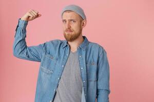 Bearded guy dressed in a denim shirt looks sad, can not boast of his strength, displeased with his physical form, raised his bent arm to demonstrate weak muscles, with chagrin looking at the camera photo