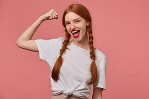 Indoor portrait of young ginger female with freckles posing over pink background showing her biceps and smiles broadly photo