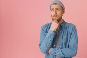 Young man is focused, keeps fist near the chin trying to remember something, looking away with sad thougtful look, ponder about something, isolated over pink background with copy space photo