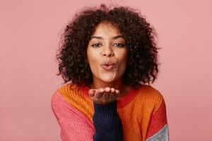 Close up portrait of charming african american girl with afro hairstyle blowing air kiss from open palm,expresses her devotion and truthful love to boyfriend, isolated on pink background photo