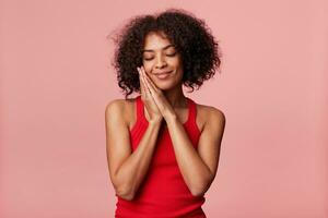 Pleased charming African American woman with afro hairstyle feels pleasure, touches her face with palm, closed eyes, rejoices, folded hands as sleepy, wearing red singlet, isolated on pink background photo