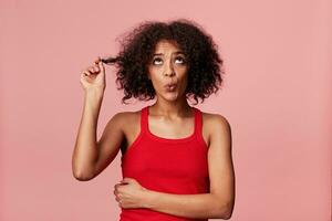 Naughty african american girl with afro hairstyle looking upwards rolled her eyes, depicts innocence, pretends not guilty,plays with stand of curly dark hair, whistles, isolated on pink background photo
