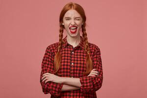 indoor shot of young redhead teenage girl, keeps her hands crossed, smiles broadly and wink with positive facial expression. Isolated over pink background photo