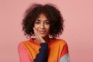 retrato de positivo satisfecho encantador africano americano niña con afro peinado mira con placer, toques su cara con palmera, mira feliz, vistiendo vistoso manga larga, aislado en rosado antecedentes foto