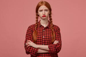 indoor shot of young redhead ginger female, wears checkered black red shirt, keeps her hands crossed, looks into camera with sad, negative facial expression, curves her lips. photo