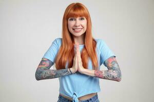 Indoor portrait of young ginger female posing over white wall pressed he palm in praying position with broad smile on her face photo