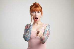 Indoor portrait of young ginger female posing over white wall points into camera with shocked facial expression photo