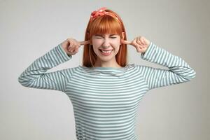 Indoor portrait of young ginger female posing over white wall plug her ears with fingers photo