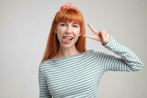 Indoor portrait of young ginger female posing over white wall showing victory sign into camera and smiles broadly photo