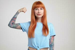 Indoor portrait of young ginger female posing over white wall showing her biceps into camera with excited facial expression photo