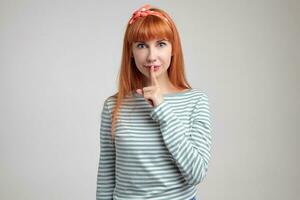 Indoor portrait of young ginger female posing over white wall showing silence gesture into camera photo