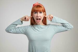 Indoor portrait of young ginger female posing over white wall plug her ears with fingers and screaming into camera photo