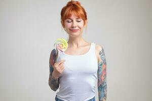 Indoor portrait of young ginger female posing over white wall holding big colorful candy and smiles photo