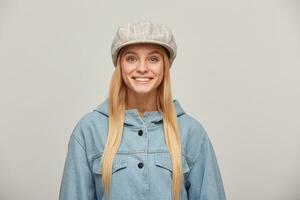 Close up of a lovely blonde young girl with long hair down, looking happy smiling, wearing oversize denim jacket and beige checked hat cap,isolated over grey background photo