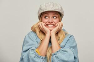 Scared blonde girl looks aside frightened afraid chattering teeth with fear,bites nails from fright, hands holding cheeks, wears oversize denim jacket and beige checked cap,on grey background photo