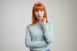 Indoor portrait of young ginger female posing over white wall looking into camera while touching her chin photo