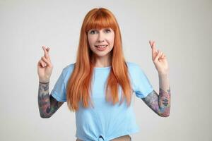 Indoor portrait of young ginger female posing over white wall crossed her fingers with excited facial expression photo