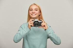 Young girl photographer looks happily smiling, holding a retro vintage photo camera in hands, in the process of shooting, working in studio, wearing blue casual sweatshirt, over grey background