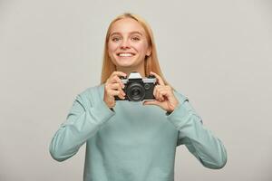 joven niña fotógrafo mira felizmente sonriente, participación un retro Clásico foto cámara en manos, en el proceso de tiroteo, trabajando en estudio, vistiendo azul casual camisa de entrenamiento, terminado gris antecedentes