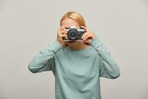 Girl covering her face with the camera making a photo session. Photographer taking photo, working in studio, in-door photography, dressed blue sweatshirt, over grey background