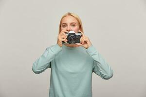 Young girl photographer looks out from behind the lens, holding a retro vintage photo camera, in the process of shooting, while working in studio, wearing blue casual sweatshirt, over grey background