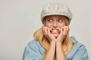 Scared blonde girl looks aside frightened afraid chattering teeth with fear,bites nails from fright, hands holding cheeks, on grey background photo