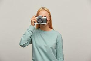 Close up of a photographer covering her face with the retro vintage camera. Happy with the camera, likes to take pictures, hand-held photography, dressed in blue sweatshirt, over grey background photo