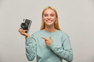 Lovely girl photographer taking a photo session, inspired by the retro vintage photo camera in hand, pointing on it with finger, dressed in blue sweatshirt, on grey background