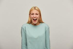 asustado rubia niña mira asustado temeroso imitar gritar gritar, pronunciar un ruidoso llamada o llorar, un expresión de un temor emoción, vistiendo azul casual sudadera,aislado terminado gris antecedentes foto