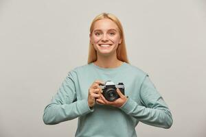 Emotional blonde photographer, looks inspired delighted, holding in front a retro vintage photo camera in hands, dressed in blue casual sweatshirt, on grey background