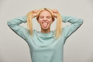 Cool young blonde girl with long hair grimacing on camera, showing tongue, eyes closed, has fun, makes two tails on her head, wearing blue casual sweatshirt,i isolated over grey background photo