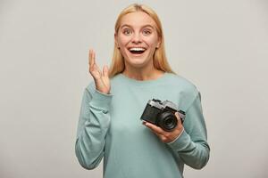 Blonde girl photographer, taking a photo session, did not expect to get so many great shots, inspired by the retro vintage photo camera in hand, dressed in blue sweatshirt, on grey background