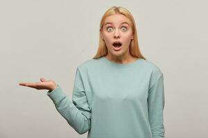 Shocked blonde young girl, looking wonder struck amazed open-eyed wide open mouth, holding the copy space on one palm, wearing blue casual sweatshirt,isolated over grey background photo