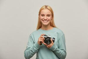 Blonde girl photographer, nicely widely smiles, looks happy delighted, holds in front a retro vintage photo camera in hands, dressed in blue sweatshirt, on grey background