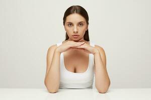 Beautiful woman with perfect healthy fresh skin sits at the table, looks at camera as in the mirror, palms folded under chin, carefully examines her face in details, not very satisfied with result photo