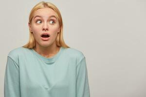 Worried scared blonde girl looks aside around with mouth opened, frightened afraid, presses down her head in shoulders, wearing blue casual sweatshirt, copy space on the right, grey background photo