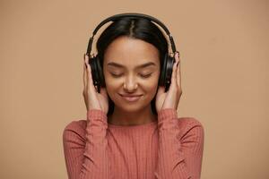 Portrait of a young mixed race girl listens to her favorite music in big black headphones, closing her eyes from pleasure, enjoyment, music lover, likes lyric songs about love, on beige background photo