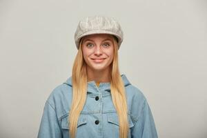 bonito encantador joven rubia niña con largo pelo abajo, mirando contento, sonriente, vistiendo demasiado grande mezclilla chaqueta y beige comprobado sombrero tapa,aislado terminado gris antecedentes foto