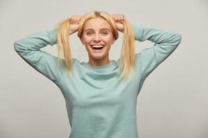 Lovely young blonde girl with long hair down has fun jokes, smiles, rejoices, makes two tails on her head, wearing blue casual sweatshirt,isolated over grey background photo