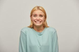 Scared blonde girl looks frightened afraid chattering teeth with fear, see something unexpected scary in front, here's to cry or scream, wearing blue casual sweatshirt,isolated over grey background photo