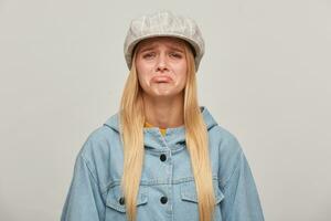 Emotional blonde girl, offended, upset, about to cry, lip pouting, did not get what she wanted, wearing oversize denim jacket and beige checked hat cap,isolated over grey background photo