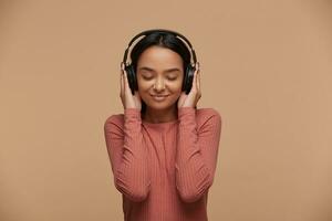 A young mixed race girl listens to her favorite music in big black headphones, closing her eyes from pleasure, enjoyment, music lover, likes lyric songs about love, on beige background photo