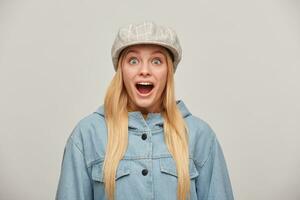 Lovely blonde girl with long hair down, looks delighted gladden amazed, mouth open, like dream came true, big discount in shop,wears oversize denim jacket and beige checked cap, over grey background photo