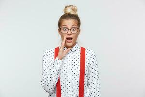 Young woman whispers secret, gossip and spread rumors. Emotional girl feels amazed to hear confidential information. Secrecy, gossiping. In a white shirt with black dots, glasses, red suspenders photo