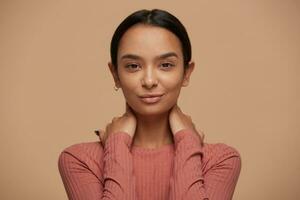 Portrait of mysterious intriguing beautiful satisfied woman, with dark hair putted back, touching her neck with both hands holding palms on neck, on beige background photo