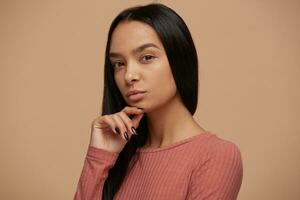 Attractive young woman looks puzzled, trying to remember something important, looks camera on keep one hand on a chin, thoughtful, wears casual clouth, on a on beige background photo